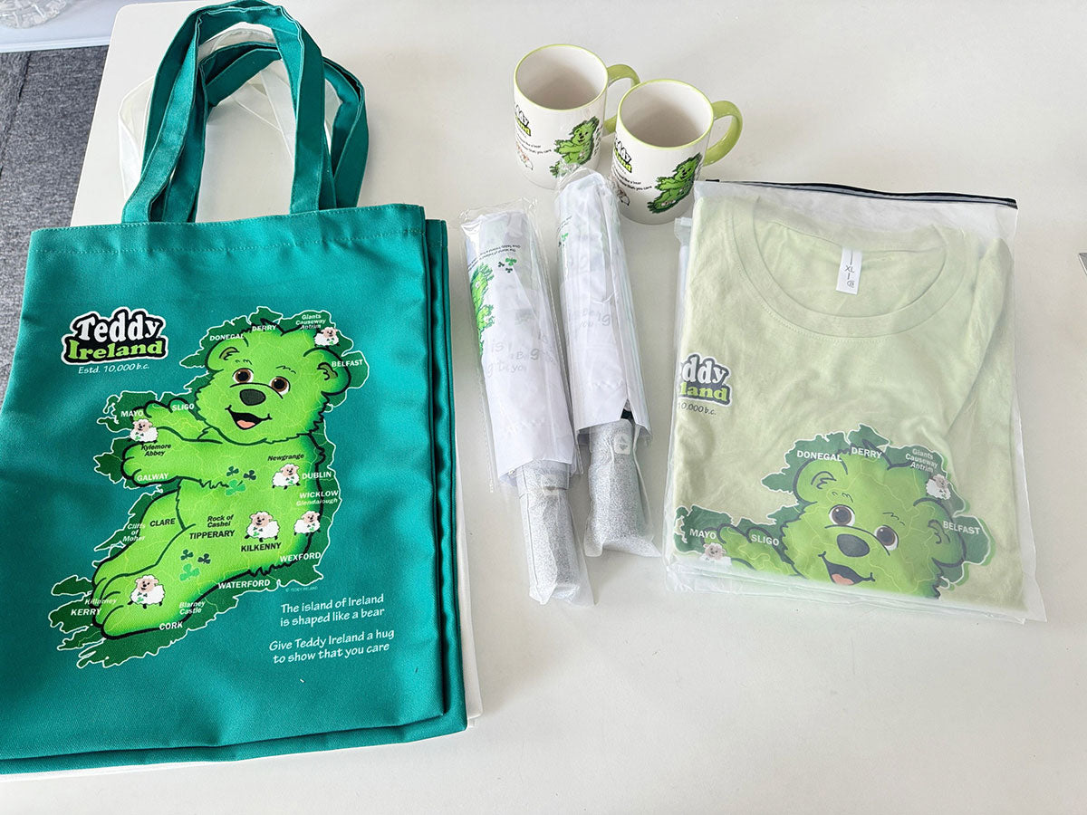 Green tote bag, t-shirt, and mugs with Teddy Ireland character bear design on a white surface